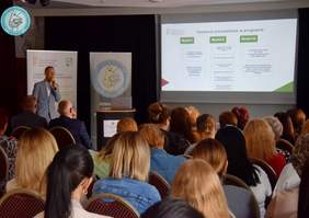 Pokaż zdjęcie: Pracownik PFRON omawia program CIDON, za nim baner z logo PFRON, w centralnej części widoczny ekran z prezentacją. W dolnej stronie zdjęcia słuchający prezentacji  goście wydarzenia. Całość odbywa się w lekkim zaciemnieniu światła.  