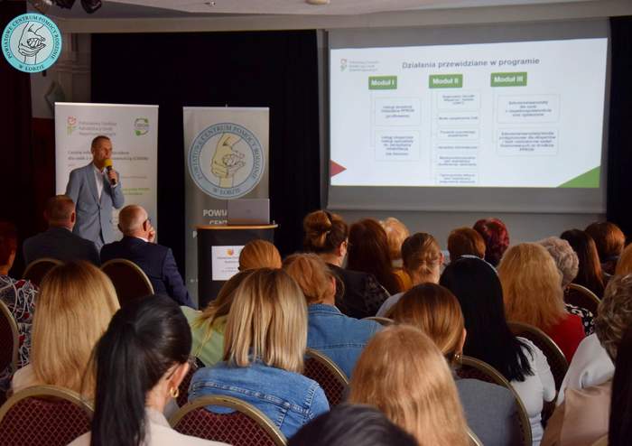 Pokaż zdjęcie: Pracownik PFRON omawia program CIDON, za nim baner z logo PFRON, w centralnej części widoczny ekran z prezentacją. W dolnej stronie zdjęcia słuchający prezentacji  goście wydarzenia. Całość odbywa się w lekkim zaciemnieniu światła.  