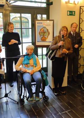 Pokaż zdjęcie: Trzy kobiety oraz mężczyzna, wśród nich kobieta na wózku inwalidzkim, tłumaczka języka migowego oraz kobieta przemawiająca do mikrofonu.