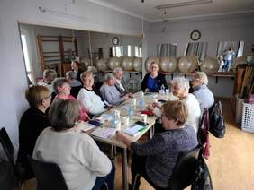 Pokaż zdjęcie: Przy prostokątnym stole siedzą kobiety w różnym wieku. Rozmawiają ze sobą i uśmiechają się do siebie. Na ścianach wiszą lustra, a na półkach leżą piłki gimnastyczne do ćwiczeń. 