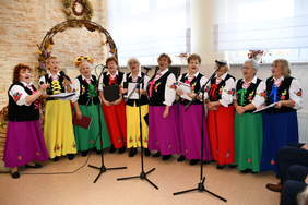 Pokaż zdjęcie: Na scenie stoją w półkolu kobiety z lokalnego zespołu ludowego. Śpiewają piosenkę. Są ubrane w długie suknie w żywych kolorach. 