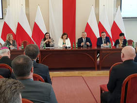 Grupa kilku osób, kobiet i mężczyzn przy stole prezydialnym, za nimi flagi biało-czerwone, widać także zebranych gi