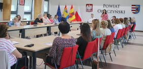 Pokaż zdjęcie: Przy stołach ustawionych w kwadrat siedzą na krzesłach ludzie. Tylko jedna pani stoi i przemawia do mikrofonu. Jest ubrana w czerwoną sukienkę w kolorowe kwiaty. 