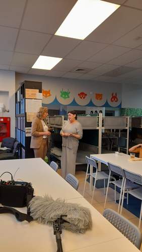 Pokaż zdjęcie: W ośrodku recepcyjnym w sali dziennej, gdzie stoją piętrowe łóżka rozmawiają dwie kobiety. Jedna z nich blondynka ubrana jest w beżowy garnitur, druga z pań ma na sobie dresy, jest mieszkanką ośrodka. Na stole leży mikron oraz futerał ze stacją nagłaśniającą.. 