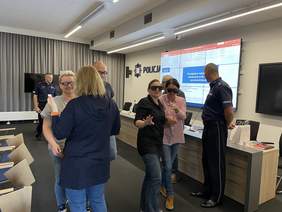 Pokaż zdjęcie: Na sali konferencyjnej kliku mężczyzn i kobiet ma założonych okulary treningowe. Wyciągają ręce do przodu co świadczy o problemach z widzeniem.