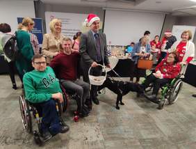 Pokaż zdjęcie: Grupa uśmiechniętych, świątecznie ubranych osób pozuje do zdjęcia. Osoby z różnymi niepełnosprawnościami.