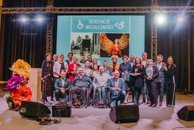 Pokaż zdjęcie: Grupa dwudziestu trzech osób stoi na scenie udekorowanej sztucznymi kwiatami. Część z tych osób trzyma w dłoniach statuetki. Na przodzie znajdują się dwaj panowie, którzy poruszają się na wózku. W tle duży ekran z napisem „Generacje Niezależności”.