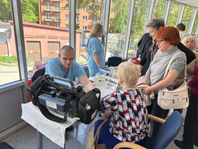 Pokaż zdjęcie: Przy stole siedzi mężczyzna w fartuchu i kobieta naprzeciw która podaje mu rękę. Obok na stole sprzęt medyczny. 