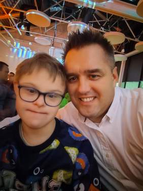 Pokaż zdjęcie: Do zdjęcia pozują dwie osoby: mężczyzna i chłopiec z Zespołem Downa. Uśmiechają się. 