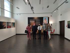 Pokaż zdjęcie: Na zdjęciu znajduje się pięć osób w dużym pomieszczeniu pomalowanym na biało. Osoby stoją w jednym rzędzie na tle dużego zdjęcia z opisem wystawy Na ścianach wiszą fotografie. 