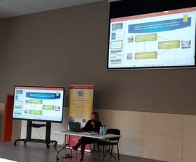 Pokaż zdjęcie: sala konferencyjna na środku stoi biurko przy którym siedzi osoba i przedstawia prezentację projektu kompleksowej rehabilitacji na górze wyświetla się prezentacja na dużym telebimie