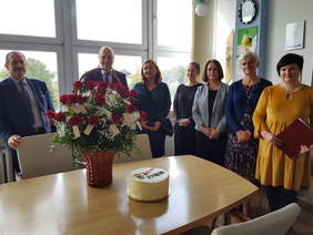 W gabinecie pod oknem stoją osoby, od lewej dwóch mężczyzn oraz 4 kobiety. Wszyscy uśmiechają się. Przed nimi na stole stoi kosz z bukietem czerwonych róż oraz okrągły tort