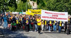 Pokaż zdjęcie: Osoby ubrane w żółte koszulki idą środkiem ulicy, w dłoniach trzymają instrumenty muzyczne bębenki, przed sobą niosą banery pierwszy z nich z napisem Marsz Godności Osób Niepełnosprawnych.