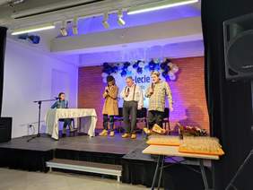 Pokaż zdjęcie: Na scenie zaaranżowanej jako urząd odbywa się scenka kabaretowa, przy biurku siedzi kobieta,  trzech mężczyzn petentów stoi przed biurkiem frontem do widowni.