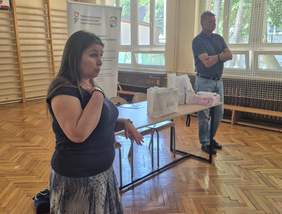 Pokaż zdjęcie: Przy stoliku ma którym stoją torby z prezentami, leżą katalogi i ulotki stoją dwie osoby, mężczyzna i kobieta. Kobieta wyraźnie gestykuluje, tłumacząc uczestnikom konferencji.