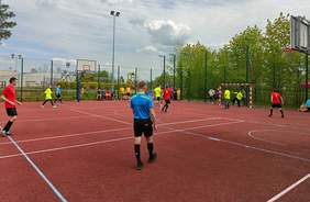 Pokaż zdjęcie: Zawodnicy w kolorowych koszulkach rozgrywają mecz piłki nożnej na otwartym boisku. W tle widać sędziów, drużyny siedzące na ławkach oraz kibiców za ogrodzeniem boiska. Po prawej stronie akcja toczy się przy bramce bronionej przez zawodnika w żółtej koszulce.