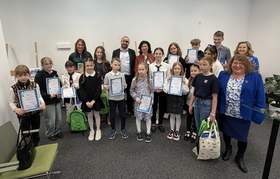 Duża grupa osób: dorośli i dzieci z dyplomami pozują do grupowego zdjęcia. W tle drewniane sztalugi z pracami plastycznymi