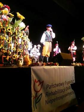 Pokaż zdjęcie: Chłopiec  w stroju ludowym gwiżdżący na gwizdku, w tle widać osoby z zespołu oraz płot opleciony słonecznikami. Na dole sceny wisi baner z logo PFRON.