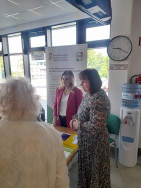 Pokaż zdjęcie: Przy stole z ulotkami stoją trzy kobiety, które rozmawiają.