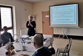 Pokaż zdjęcie: Kobieta stoi na środku sali, omawia wyświetlaną na ścianie prezentację. Słuchacze siedzą przy stole.