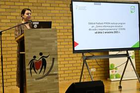 Pokaż zdjęcie: Kobieta stoi na scenie za mównicą z mikrofonem i prowadzi prezentacje na temat „Wsparcie programowe dedykowane osobom z niepełnosprawnościami”.  W tle widoczny ekran z wyświetlana prezentacją.