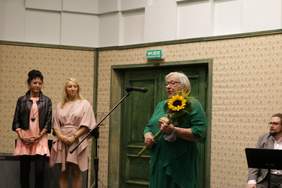 Pokaż zdjęcie: Przed mikrofonem stoi starsza kobieta ubrana w zielony kostium. Na nosie ma oku. W ręku trzyma kwiaty. Z lewej strony zdjęcia stoją dwie kobiety ubrane w jasne sukienki. Jedna z kobiet ma ubraną jeszcze czarną skurzaną kurtkę.