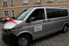 Pokaż zdjęcie: Srebrny mikrobus z naklejką informującą, że pojazd przystosowany jest do przewozu osób z niepełnosprawnością. W tle fragment budynku oraz grupa osób poruszających się na wózkach.