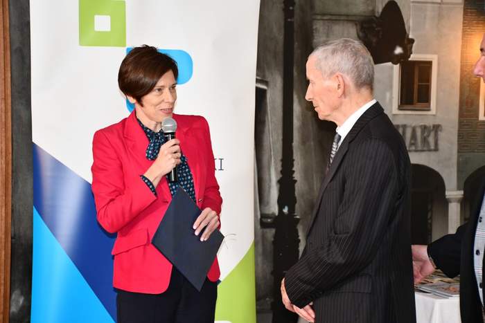 Pokaż zdjęcie: Po lewej stronie, na tle banera powiatu poznańskiego stoi kobieta ubrana w czerwoną marynarkę, która trzyma w jednej ręce mikrofon, a w drugiej teczkę z listem gratulacyjnym. Po prawej stronie stoi starszy mężczyzna w garniturze.