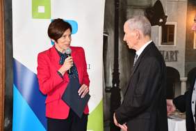 Pokaż zdjęcie: Po lewej stronie, na tle banera powiatu poznańskiego stoi kobieta ubrana w czerwoną marynarkę, która trzyma w jednej ręce mikrofon, a w drugiej teczkę z listem gratulacyjnym. Po prawej stronie stoi starszy mężczyzna w garniturze.