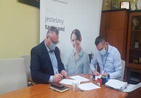 Pokaż zdjęcie: pomieszczenie Oddziału Łódzkiego PFRON dwóch mężczyzn siedzi przy stole i podpisuje dokumenty dotyczące dofinansowania ze środków PFRON