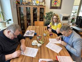 Pokaż zdjęcie: Przy drewnianym dużym stole siedzą trzy osoby, kobieta w blond włosach, kolorowym sweterku i dwaj mężczyźni. Jeden w swetrze drugi w jasnej marynarce, niebieskiej koszuli. Panowie podpisują dokumenty. Kobieta uśmiecha się do zdjęcia Za nimi komo