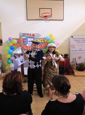 Pokaż zdjęcie: występ artystyczny odbywa się na sali gimnastycznej, dwóch uczestników WTZ ubranych w kolrowe stroje przedstawia scenkę humorystycznego scenariusza. Na drugim planie osoby siedzące i oklaskujące aktorów 