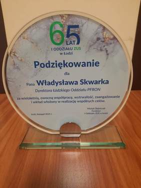 Pokaż zdjęcie: Na stole stoi statuetka z podziękowaniami za wieloletnią współpracę dla dyrektora Łódzkiego Oddziału PFRON. 