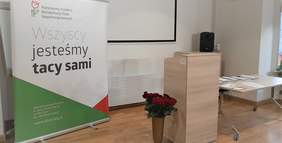 Pokaż zdjęcie: Plakat PFROn z hasłem "Wszyscy jesteśmy tacy sami", obok mównicy stoi bukiet czerwonych róż w wazonie, z drugiej strony mównicy stół z teczkami PFRON - dla laureatów konkursu
