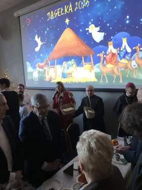 Pokaż zdjęcie: Przy wspólnym stole stoją ludzie. Składają sobie świąteczne życzenia. Wszyscy są pięknie i elegancko ubrani. 