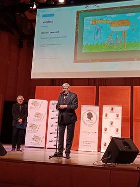 Pokaż zdjęcie: Na scenie Akademii Muzycznej stoi mężczyzna, który przemawia do mikrofonu. Obok niego stoi drugi pan. Nad nimi wisi tablica multimedialna, na której wyświetlane są nagrodzone prace plastyczne. 
