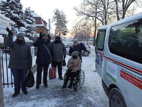 Pokaż zdjęcie: na chodniku stoją 4 osoby jedna jest na wózku inwalidzkim z boku fragment białego busa w tle ośnieżone choinki i drzewa oraz fragmenty budynków i samochodów
