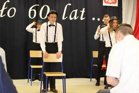 Pokaż zdjęcie: Trzech chłopców i jedna dziewczyna stoją pzrd krzesłami podczas części artystycznej. Ubrani sa w białe koszule,czarne spodnie ,na głowie mają czarne meloniki.  tle sceny napis 60 lat