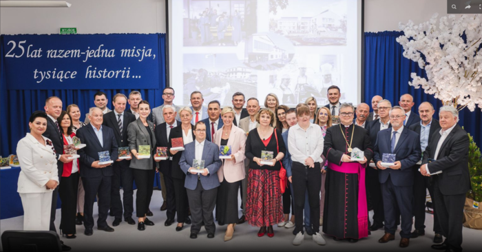 Pokaż zdjęcie: Alt tekst (do 250 znaków):  Grupowe zdjęcie uczestników jubileuszu 25-lecia Fundacji „Uśmiech Dziecka” w Szklanej. W centrum osoby trzymające książki, obecny także duchowny w stroju liturgicznym. Tło z napisem i projekcją.  