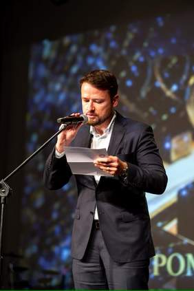 Pokaż zdjęcie: Dyrektor Oddziału Zachodniopomorskiego PFRON - w garniturze czyta tekst z kartki, stojąc przy mikrofonie na scenie podczas gali. W tle widoczna rozmyta projekcja ze złotymi i niebieskimi wzorami.