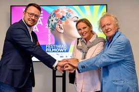 Mężczyzna, kobieta i starszy mężczyzna stoją razem przed ekranem z grafiką 23. Europejskiego Festiwalu Filmowego Integracja „Ja i Ty”. Trzy osoby symbolicznie kładą dłonie jedna na drugiej.