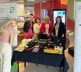 Pokaż zdjęcie: Zdjęcie stolika na którym leżą tablet, aparat, tablet, laptop, pętla indukcyjna, powiększalnik i ulotki. Za stołem stoją cztery kobiety. Po obydwu stronach stolika stoją rollupy.