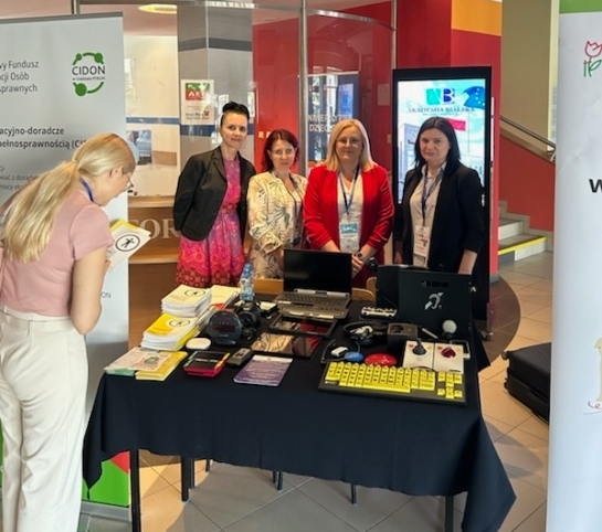 Pokaż zdjęcie: Zdjęcie stolika na którym leżą tablet, aparat, tablet, laptop, pętla indukcyjna, powiększalnik i ulotki. Za stołem stoją cztery kobiety. Po obydwu stronach stolika stoją rollupy.