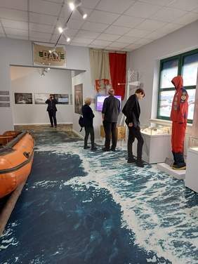 Pokaż zdjęcie: Sala poazowa w muzeum, na podłodze jest zdjęcie wody i fal, po lewej stronie widac pomarańczowy ponton, po prawej stronie stoją 3 osoby, oglądają eksponaty znajdujące się w gablotach. Na ścianie zamontowany jest telewizor z projekcja multimedialną.