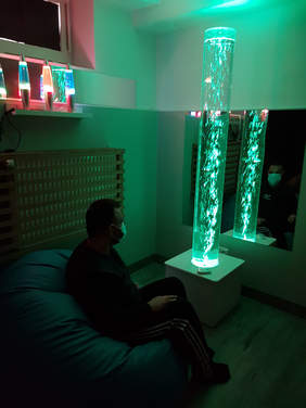 Pokaż zdjęcie: W ciemnym pokoju na miękkim siedzisku siedzi mężczyzna w maseczce, przed nim na niskiej białej szafce stoi przezroczysta zielona, podświetlona tuba w której pływają świetlne niteczki. Za tubą na ścianie jest lustro, w którym odbija się tuba o