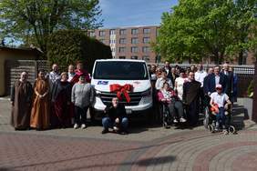 Pokaż zdjęcie: Duża grupa osób, w tym osoby z niepełnosprawnościami oraz ich opiekunowie, pozuje do wspólnego zdjęcia przed białym samochodem Caritas z czerwonymi kokardami i oznaczeniem dla osób niepełnosprawnych. W tle drzewa, budynki mieszkalne i jasne niebo.