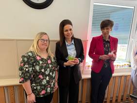 Pokaż zdjęcie: Zdjęcie trzech kobiet stojących obok siebie w pomieszczeniu, przy ścianie i oknie. Laureatka konkursu stoi pierwsza od lewej i ma blond włosy do ramion, okulary i bluzkę w ciemnym kolorze we wzór w jasnych barwach. Stoi i patrzy w stronę aparatu. Kobieta w środku ma ciemne włosy upięte z lewej strony, czarną marynarkę i jasnoniebieską koszulę trzymając w dłoniach zielone serce. Kobieta po prawej ma krótkie ciemne włosy, czerwoną marynarkę i ciemną bluzkę. Trzyma w dłoniach czerwone serce. W tle jest jasna ściana z częściowo widocznym zegarem u góry oraz okno z roletą. W dolnej części ściany widać zabudowany drewnianymi listewkami grzejnik.