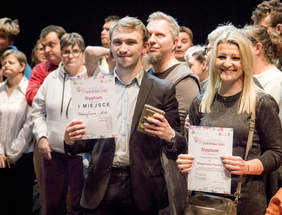Pokaż zdjęcie: Na tle dużej grupy osób młody mężczyzna ubrany w garnitur oraz kobieta blondynka pozują z dyplomami do pamiątkowego zdjęcia.