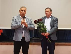 Pokaż zdjęcie: Dwóch mężczyzn stoi ubranych w marynarkach. Jeden trzyma w ręku mikrofon i mówi drugi trzyma w ręku czerwone róże.