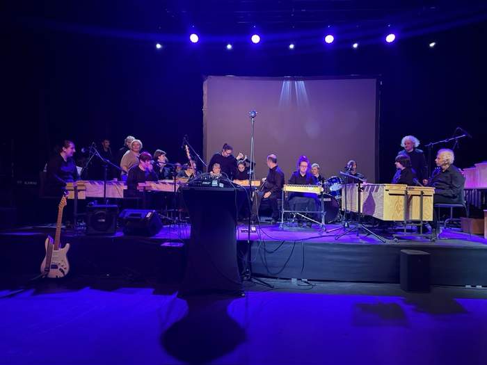 Pokaż zdjęcie: Na zdjęciu widać orkiestrę perkusyjną Gamelan z PSONI Szczecin podczas występu scenicznego. Grupa składa się z kilkunastu muzyków w różnym wieku, siedzących przy tradycyjnych instrumentach perkusyjnych ustawionych w półokręgu na podświetlonej scenie. W centralnym punkcie znajduje się wolnostojący mikrofon, a przed nim — stół z konsoletą dźwiękową. W tle widać ciemne kurtyny i duży ekran bez wyświetlanej treści. Scena oświetlona jest niebiesko-fioletowym światłem, które tworzy nastrojowy klimat. Po lewej stronie oparta jest elektryczna gitara, a po prawej – widoczna jest perkusja marki Yamaha. Artyści są skupieni i przygotowani do gry, co tworzy atmosferę oczekiwania na rozpoczęcie koncertu.